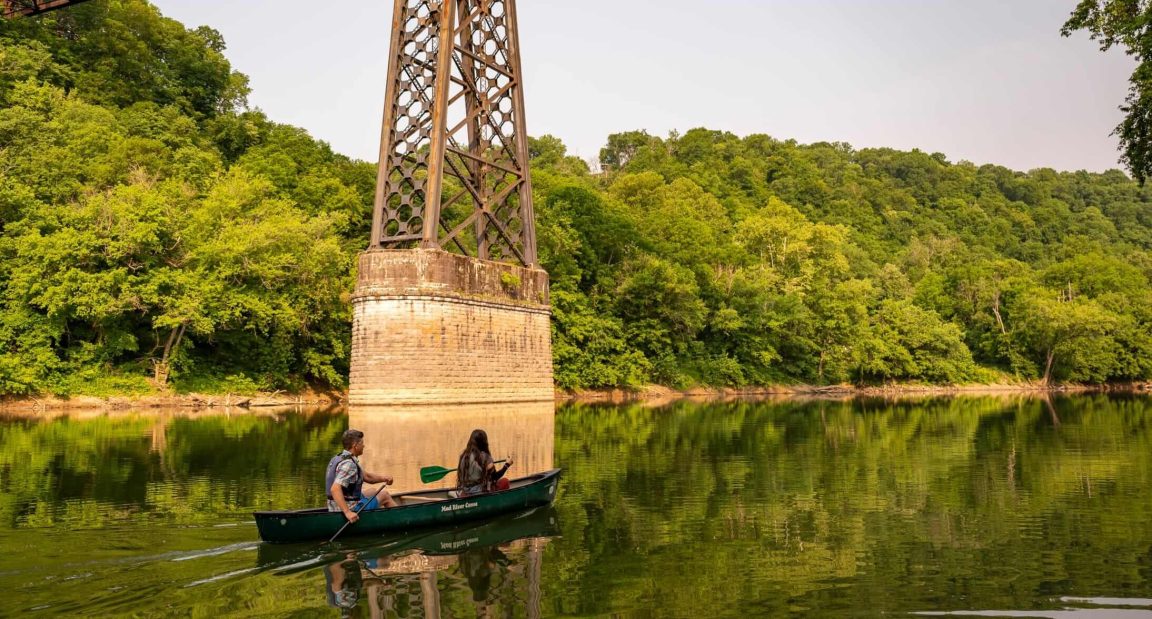 A man and a woman paddle a canoe under a bridge on a river surrounded by trees.